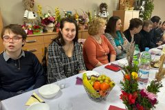 Uczestnicy podczas Spotkania Wielkanocnego siedzą przy stole, Dyrektor ŚDS dzieli się jajeczkiem