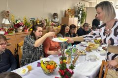 Uczestnicy podczas Spotkania Wielkanocnego siedzą przy stole, Dyrektor ŚDS dzieli się jajeczkiem